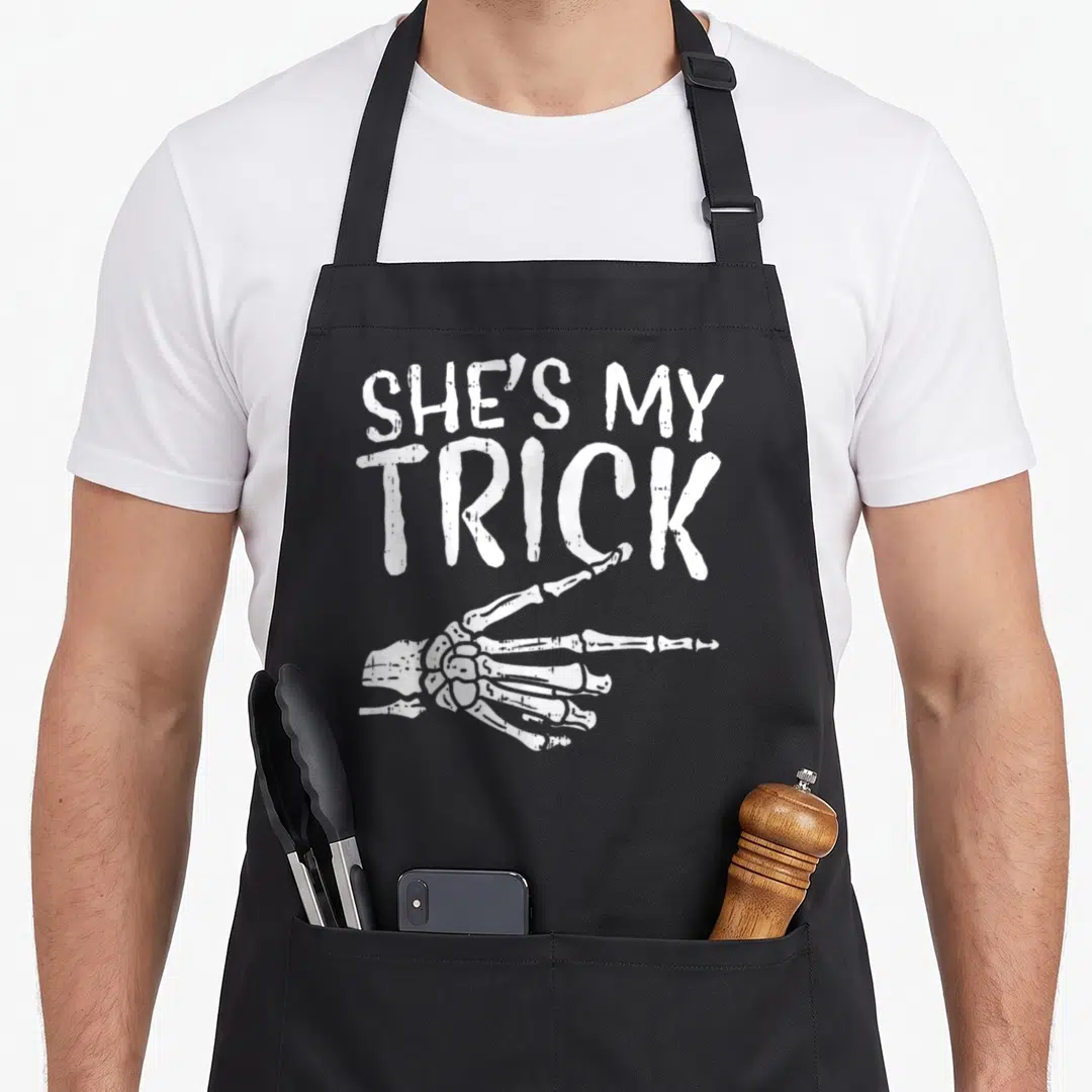 Boyfriend halloween apron - White distressed SHE'S MY TRICK text above a pointing skeleton hand — shown on a black cotton canvas apron against a white background