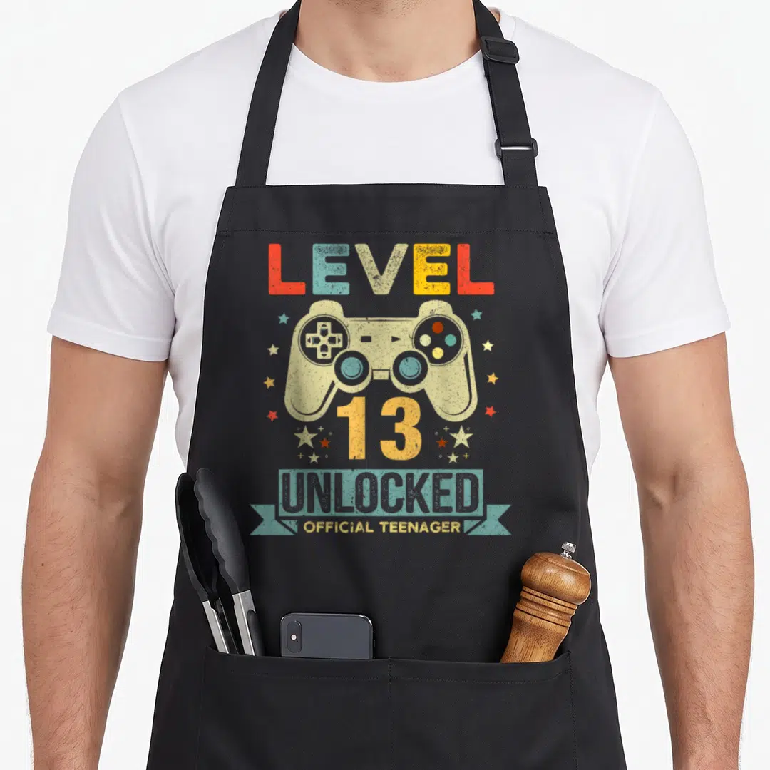 Unlocked 13th gamer birthday apron - Retro game controller surrounded by stars with LEVEL 13 UNLOCKED OFFICIAL TEENAGER — shown on a black cotton canvas apron against a white background