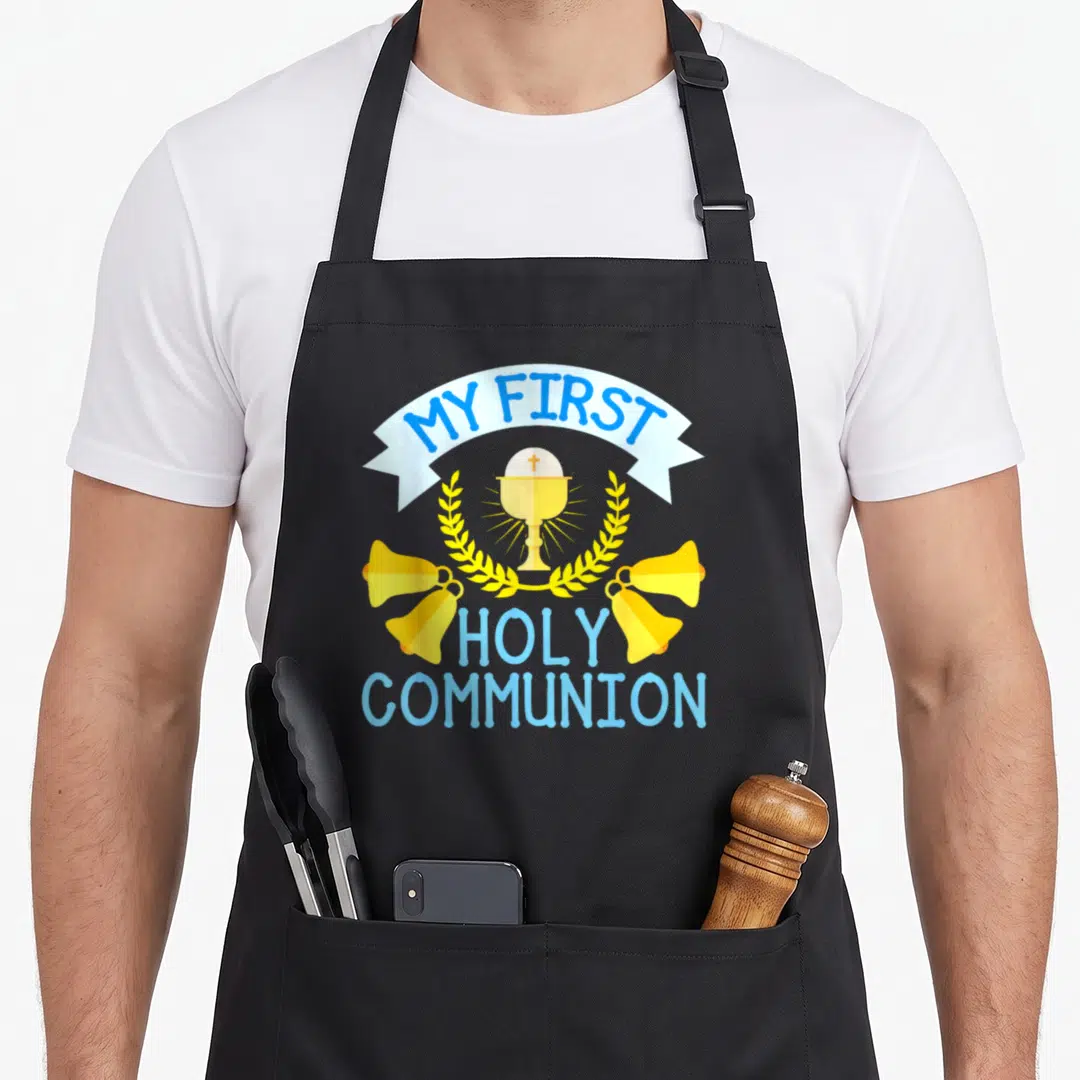 Cross communion aprons - Cartoon chalice host bells and My First Holy Communion text in blue and yellow — shown on a black cotton canvas apron against a white background