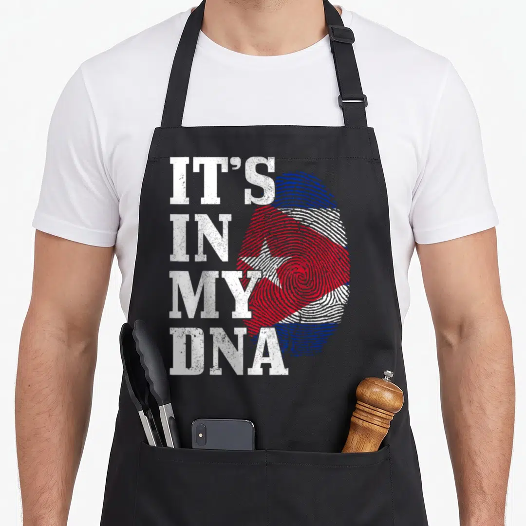 Funy pride apron - Distressed white IT'S IN MY DNA text next to a fingerprint filled with the red white and blue Cuban — shown on a black cotton canvas apron against a white background