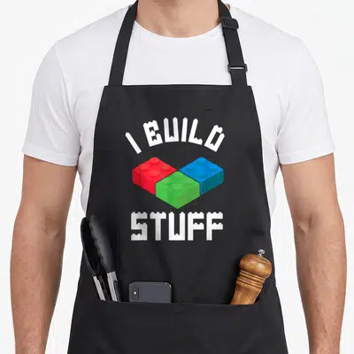 Bricklayer block & apron - White I BUILD STUFF text over red green blue interlocking toy blocks — shown on a black cotton canvas apron against a white background