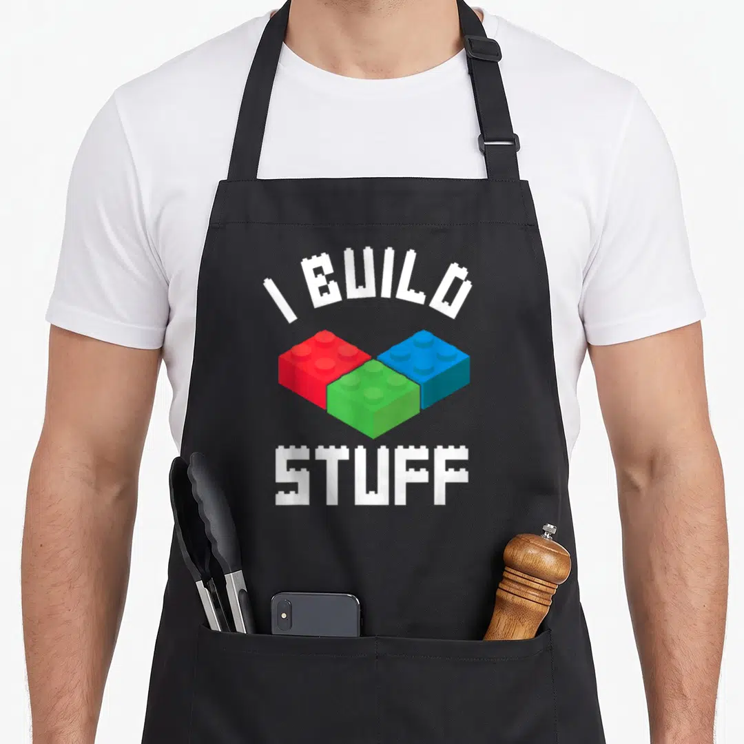 Bricklayer block & apron - White I BUILD STUFF text over red green blue interlocking toy blocks — shown on a black cotton canvas apron against a white background