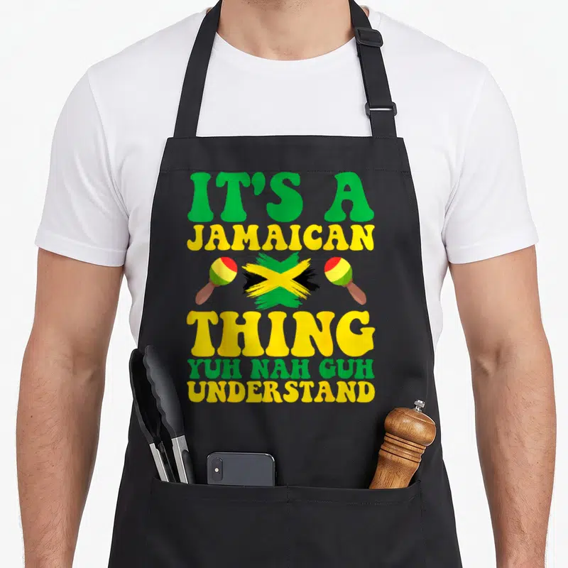 Yuh jamaican apron - Stacked IT'S A JAMAICAN THING text with brush-stroke flag and two maracas in green yellow black — shown on a black cotton canvas apron against a white background