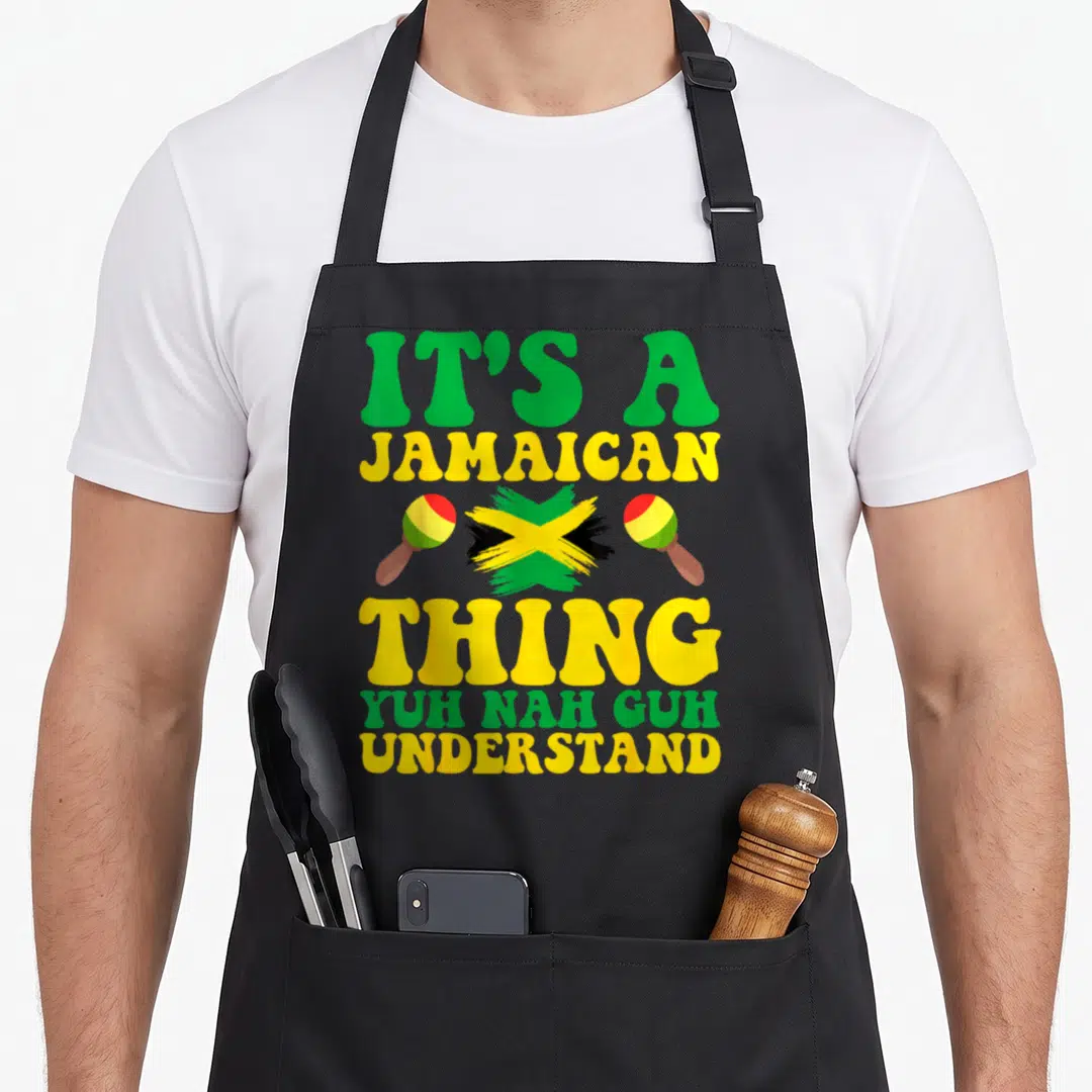 Yuh jamaican apron - Stacked IT'S A JAMAICAN THING text with brush-stroke flag and two maracas in green yellow black — shown on a black cotton canvas apron against a white background