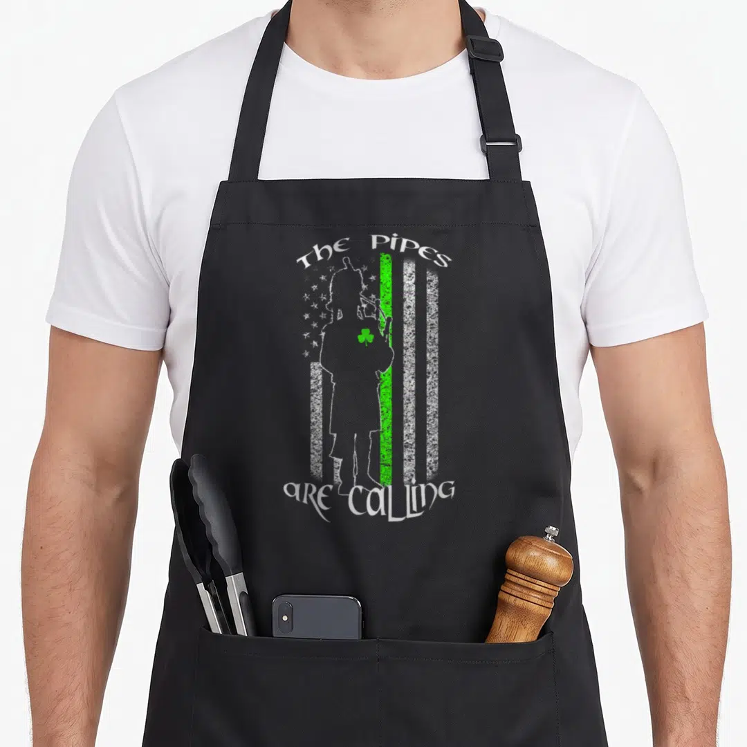 Calling pipe apron - Distressed bagpiper silhouette with green stripe and arched text The Pipes Are Calling — shown on a black cotton canvas apron against a white background
