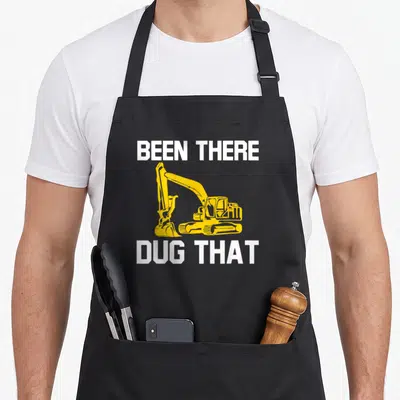 Dug heavy canvas aprons - Yellow excavator graphic between white BEEN THERE DUG THAT text — shown on a black cotton canvas apron against a white background