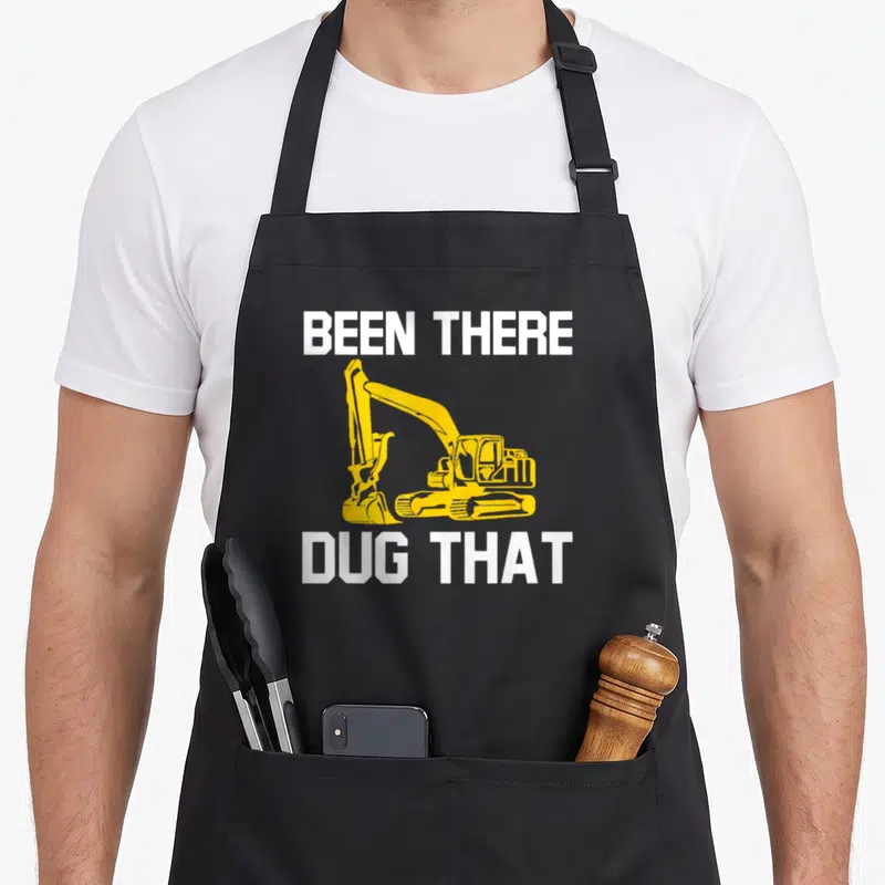Dug heavy canvas aprons - Yellow excavator graphic between white BEEN THERE DUG THAT text — shown on a black cotton canvas apron against a white background