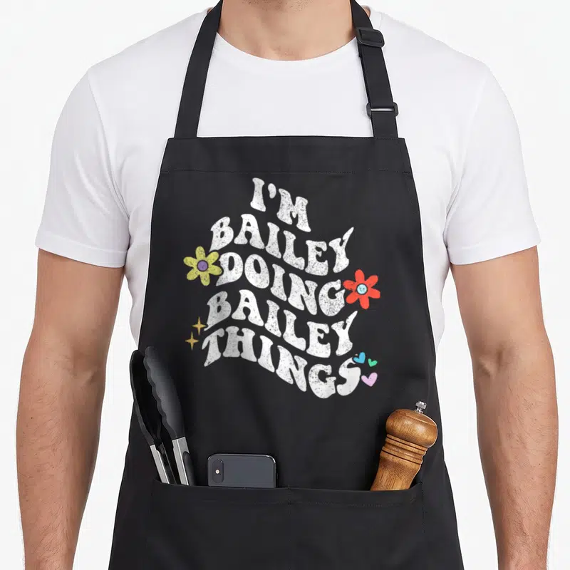 Groovy mother's apron - Wavy white text I'M BAILEY DOING BAILEY THINGS with yellow red flowers stars and hearts — shown on a black cotton canvas apron against a white background
