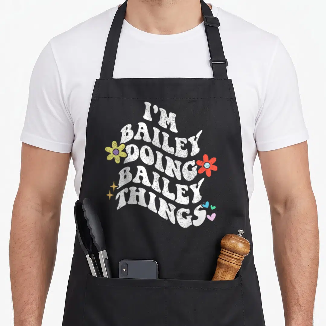 Groovy mother's apron - Wavy white text I'M BAILEY DOING BAILEY THINGS with yellow red flowers stars and hearts — shown on a black cotton canvas apron against a white background