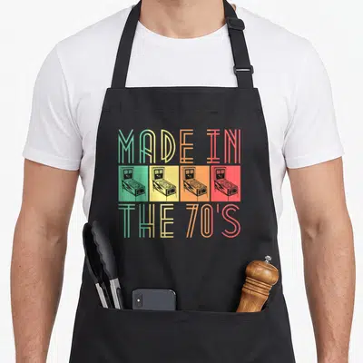 Pinball 70s apron - MADE IN THE 70'S text above and below four arcade machines on green, yellow, orange, red squares — shown on a black cotton canvas apron against a white background