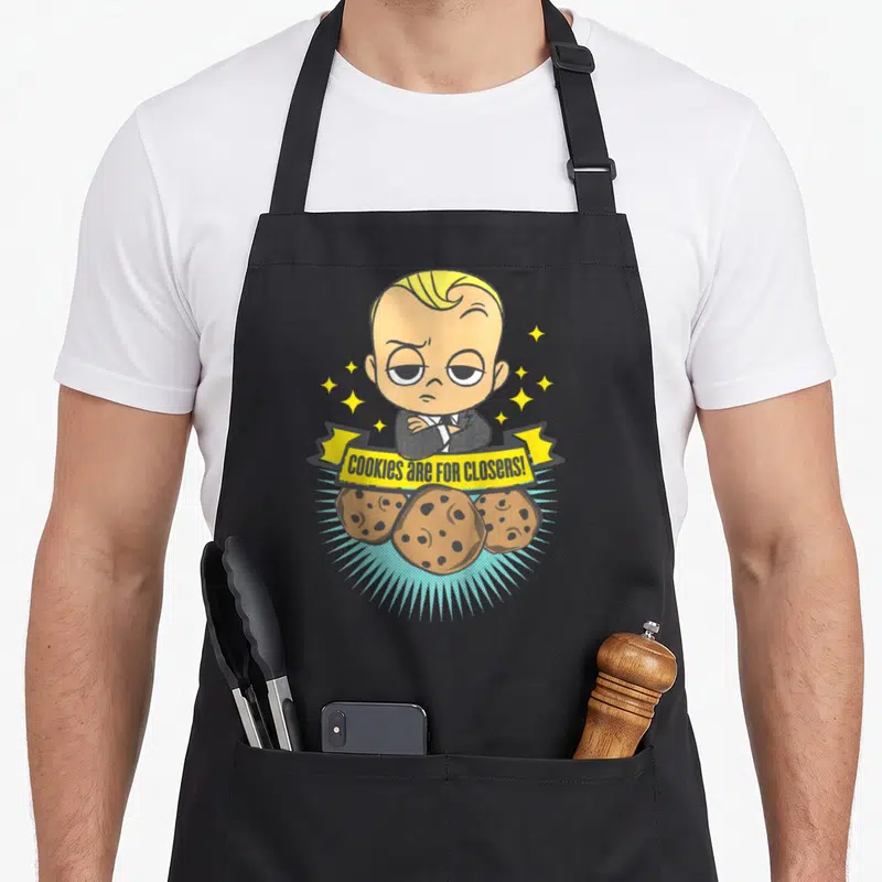 Closer cookies apron - Suited baby holding yellow ribbon with text above three cookies and stars — shown on a black cotton canvas apron against a white background