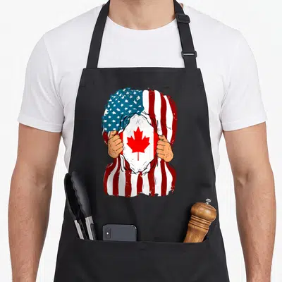 Canada inside american flag apron - Two hands tear a red white and blue USA flag to reveal a Canadian flag — shown on a black cotton canvas apron against a white background