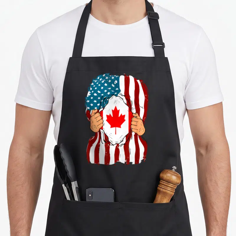 Canada inside american flag apron - Two hands tear a red white and blue USA flag to reveal a Canadian flag — shown on a black cotton canvas apron against a white background