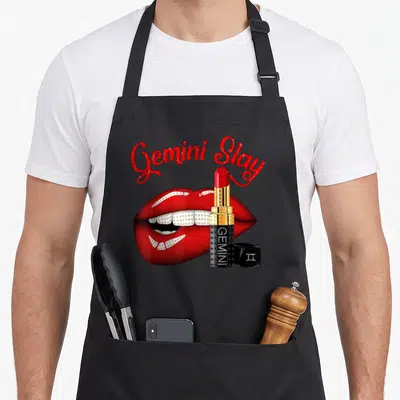 Gemini slay birthday apron - Red lips with diamond grills and gold lipstick tube under arched text — shown on a black cotton canvas apron against a white background