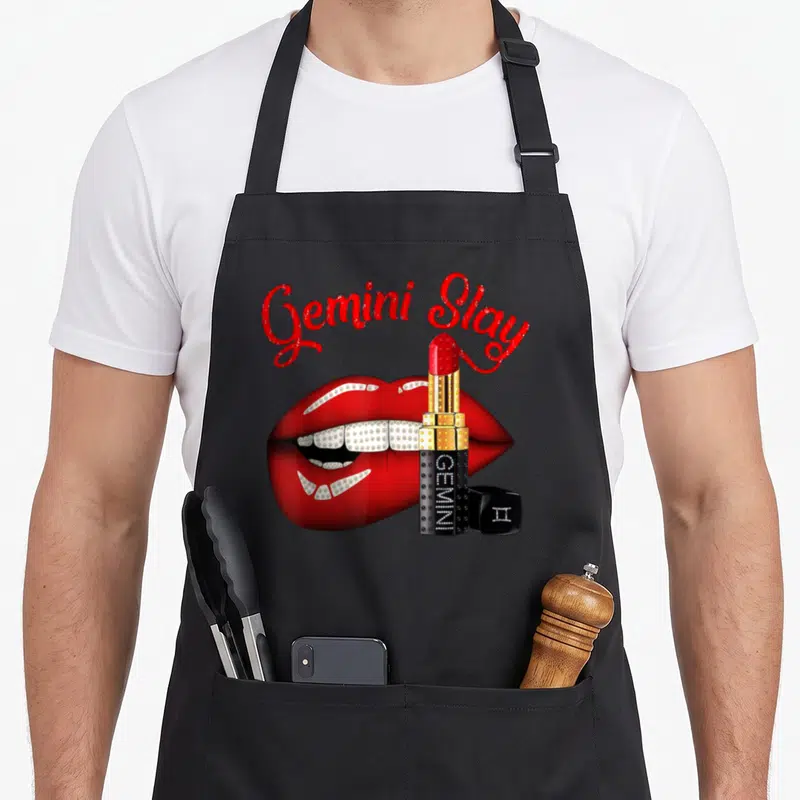 Gemini slay birthday apron - Red lips with diamond grills and gold lipstick tube under arched text — shown on a black cotton canvas apron against a white background
