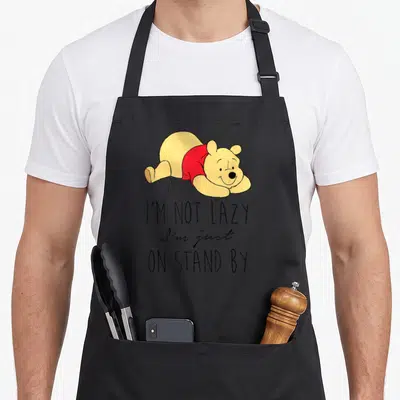 Cute lazy apron - Winnie the Pooh lying on stomach above I'M NOT LAZY I'm just ON STAND BY text — shown on a black cotton canvas apron against a white background
