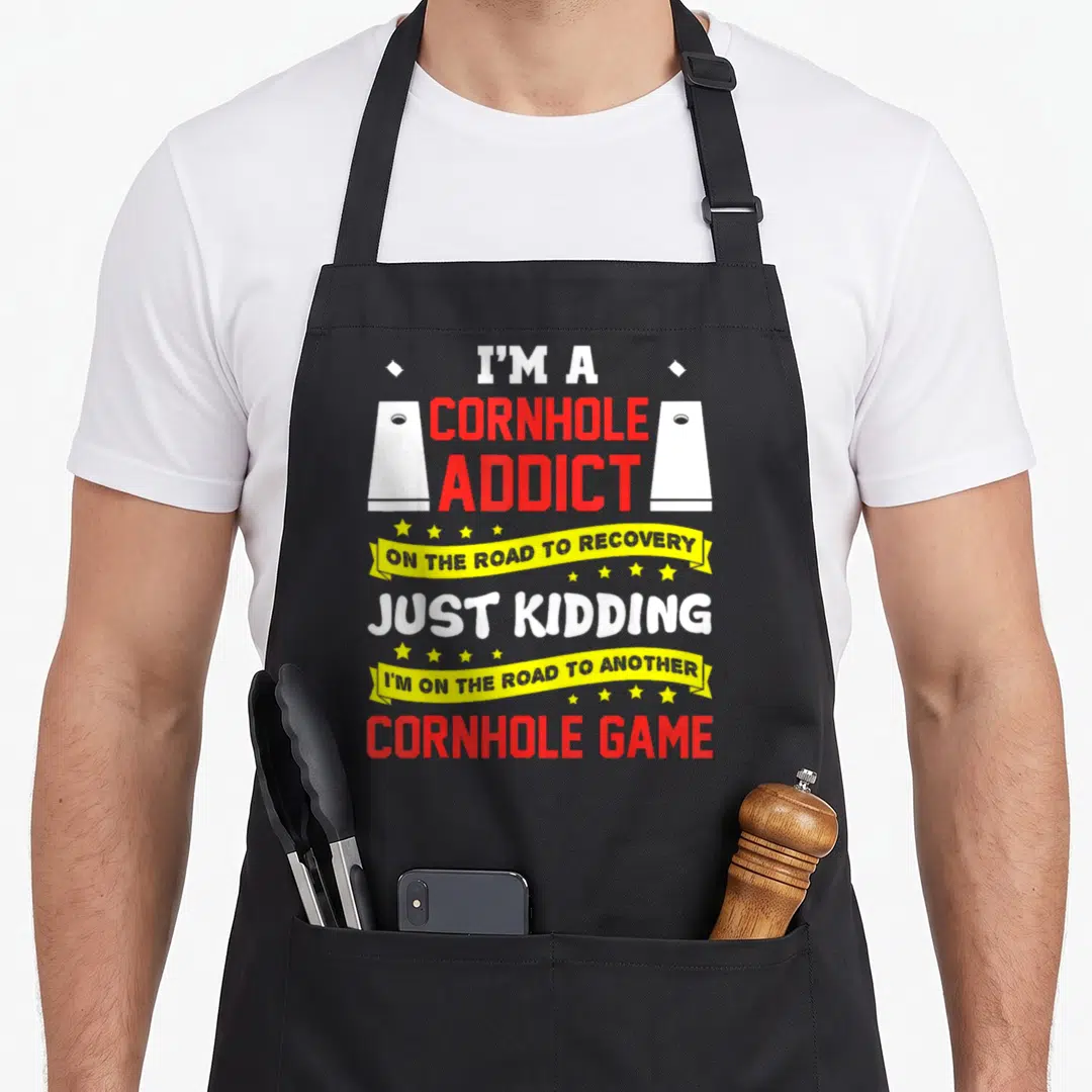 Cornhole addict game aprons - Red and white text with two white cornhole boards, yellow ribbons, and scattered stars — shown on a black cotton canvas apron against a white background
