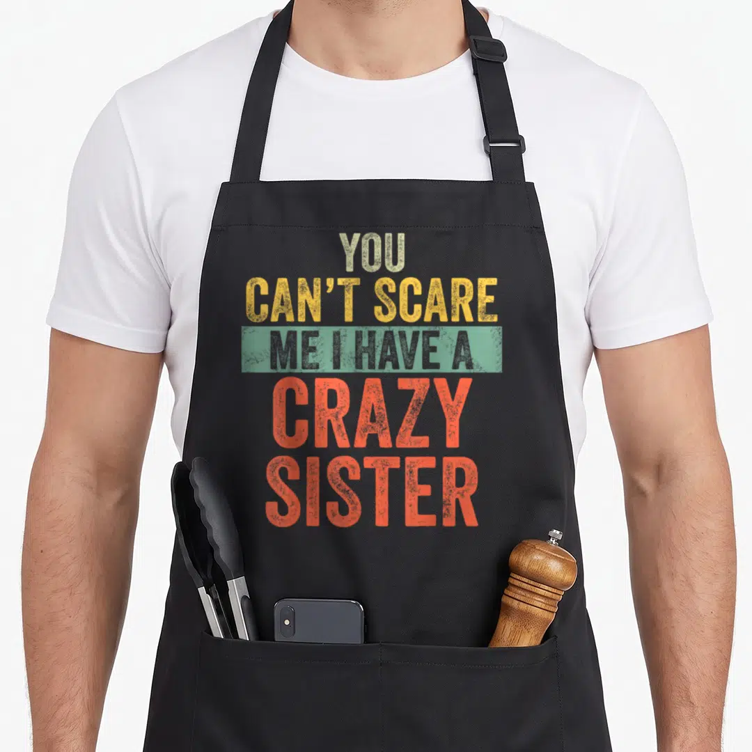 Scare joke apron - Stacked distressed text You Can't Scare Me I Have A Crazy Sister in beige yellow grey and orange red — shown on a black cotton canvas apron against a white background