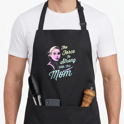 Padme strong mom apron - Distressed portrait of a woman with stacked pastel text reading The Force Is Strong With This — shown on a black cotton canvas apron against a white background
