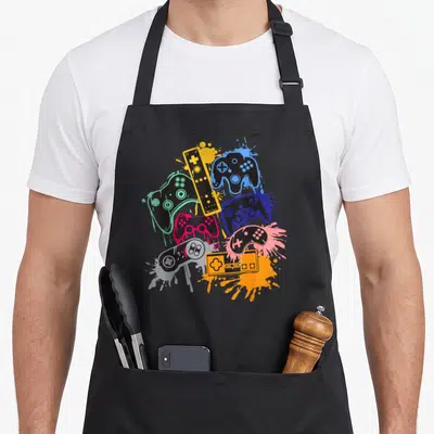 Control video game apron - Six black game controllers over yellow blue green pink grey paint splatters cartoon style — shown on a black cotton canvas apron against a white background