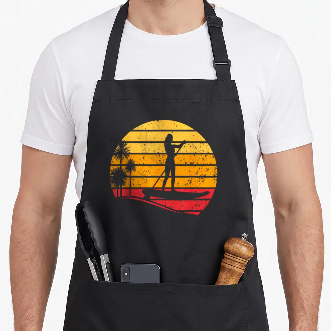 Sunset paddle apron - Grey paddleboarder silhouette on striped yellow orange red sun with palm trees — shown on a black cotton canvas apron against a white background