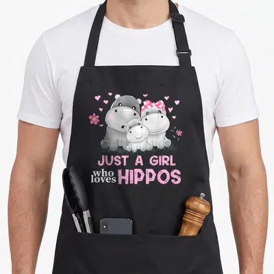 Hippo family aprons - Three cartoon hippos in grey and pink with hearts and flowers under stacked text — shown on a black cotton canvas apron against a white background