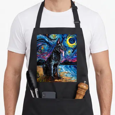 Gsd dog apron - Black German Shepherd sitting on beach under swirling blue yellow starry night sky — shown on a black cotton canvas apron against a white background