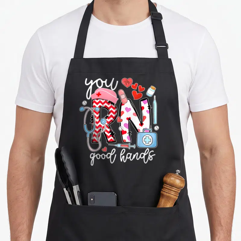 Hand nursing student apron - Cartoon RN scrubs text surrounded by stethoscope, hearts, and first aid kit in red pink — shown on a black cotton canvas apron against a white background