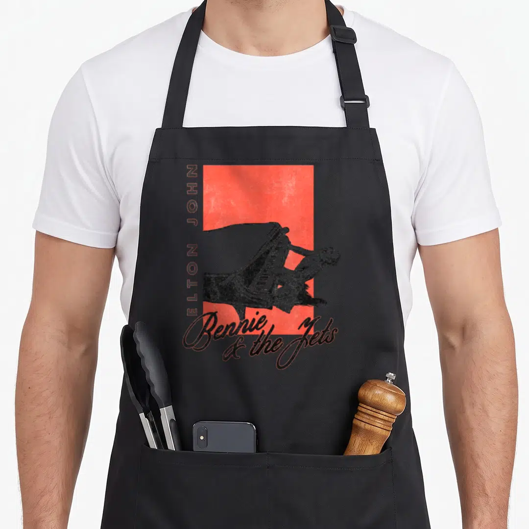 Elton piano apron - Man playing piano over red rectangle with vertical Elton John text and script Bennie and the Jets — shown on a black cotton canvas apron against a white background