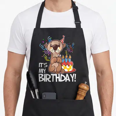 Otter birthday apron - Cartoon brown otter with party hat and blower next to cake with IT'S MY BIRTHDAY text — shown on a black cotton canvas apron against a white background