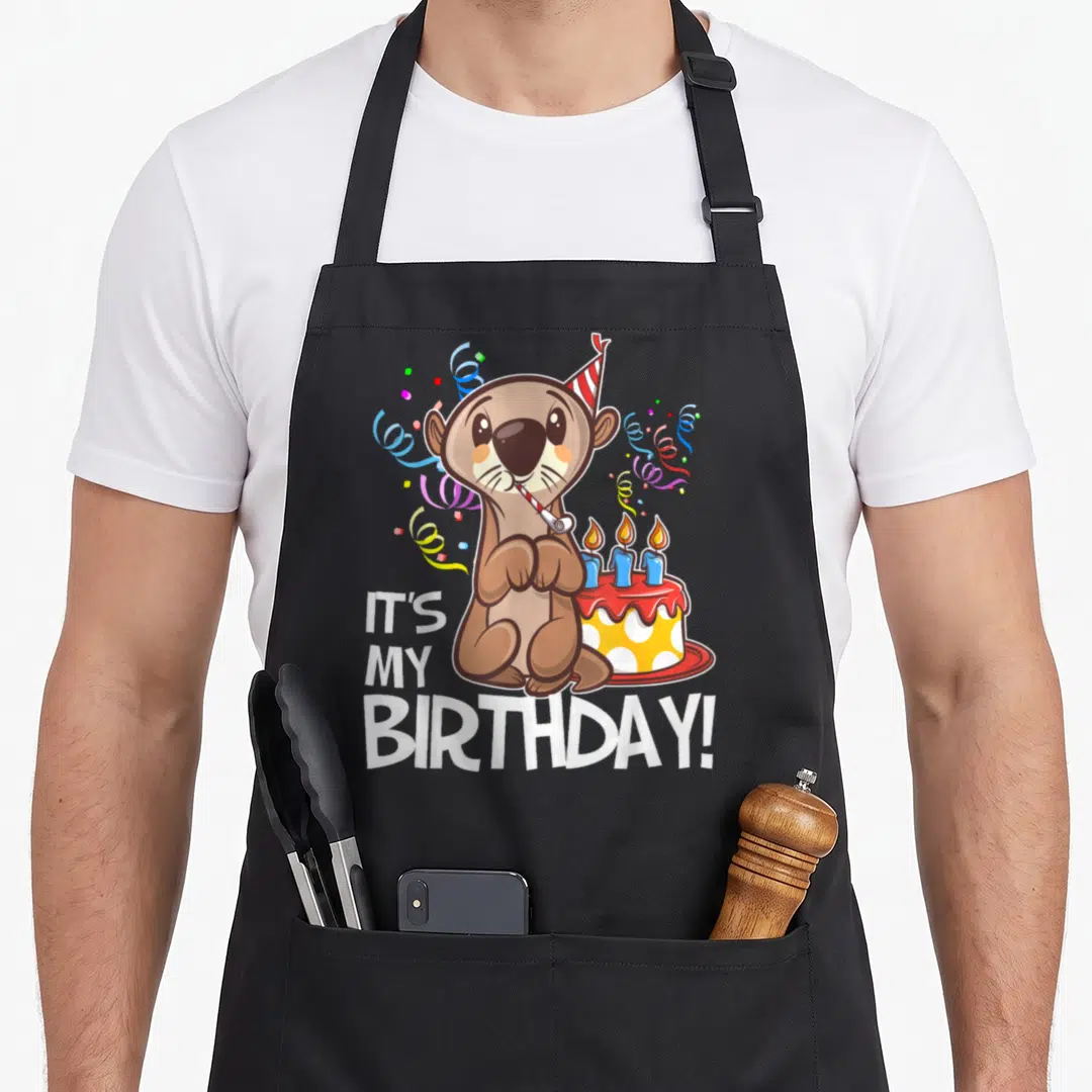 Otter birthday apron - Cartoon brown otter with party hat and blower next to cake with IT'S MY BIRTHDAY text — shown on a black cotton canvas apron against a white background
