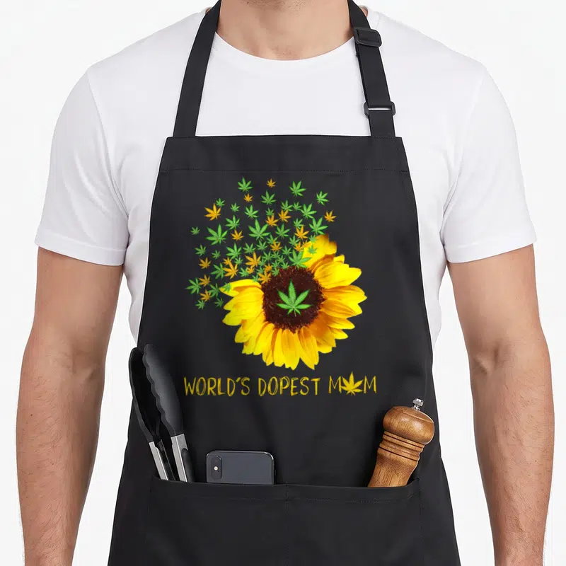 Dopest sunflower apron - Yellow sunflower with green leaf center and floating leaves above WORLD'S DOPEST MOM text — shown on a black cotton canvas apron against a white background