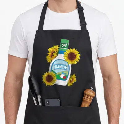 Foodie dressing apron - Cartoon ranch bottle with OPE RANCH text and four yellow sunflowers — shown on a black cotton canvas apron against a white background