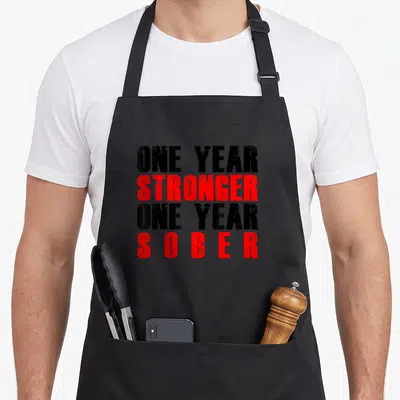 Stronger sobriety apron - Stacked black and red text reading ONE YEAR STRONGER ONE YEAR SOBER — shown on a black cotton canvas apron against a white background