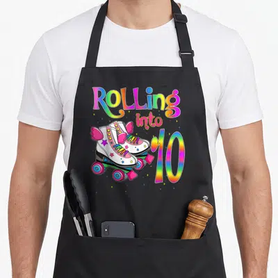 Rolling turning apron - Rainbow text Rolling into 10 above white roller skates with pink wings and stars — shown on a black cotton canvas apron against a white background