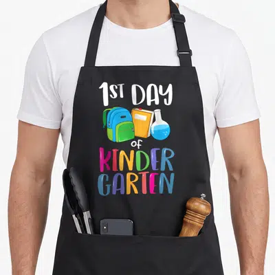 1st kindergarten back apron - Cartoon backpack, orange book, and blue flask below 1ST DAY of KINDER GARTEN text — shown on a black cotton canvas apron against a white background