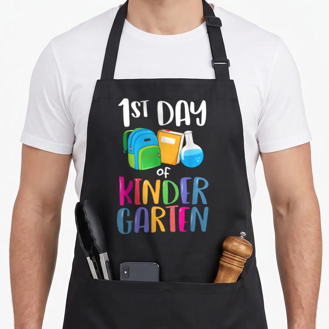 1st kindergarten back apron - Cartoon backpack, orange book, and blue flask below 1ST DAY of KINDER GARTEN text — shown on a black cotton canvas apron against a white background