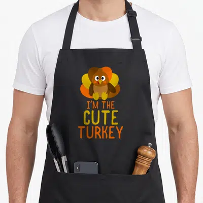 Funny family thanksgiving aprons - Cartoon turkey above stacked I'm The Cute Turkey text in autumn colors — shown on a black cotton canvas apron against a white background