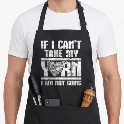 Yarn crocheting knitting apron - Distressed white text reading IF I CAN'T TAKE MY YORN I AM NOT GOING with yarn ball — shown on a black cotton canvas apron against a white background