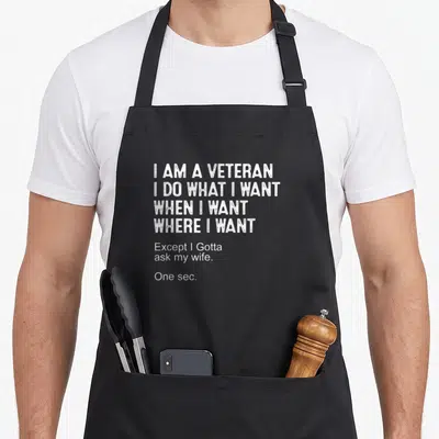 Sarcasm father day apron - White stacked typography reading I am a veteran except I gotta ask my wife on black fabric — shown on a black cotton canvas apron against a white background
