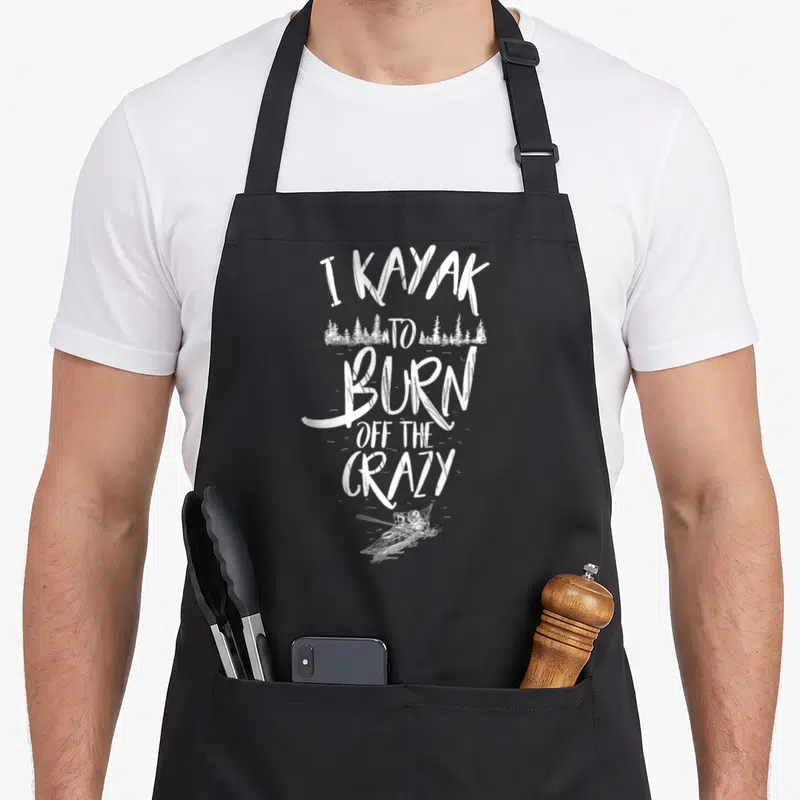 Kayaker kayak girl apron - White distressed text I KAYAK TO BURN OFF THE CRAZY with forest and kayaker silhouette — shown on a black cotton canvas apron against a white background