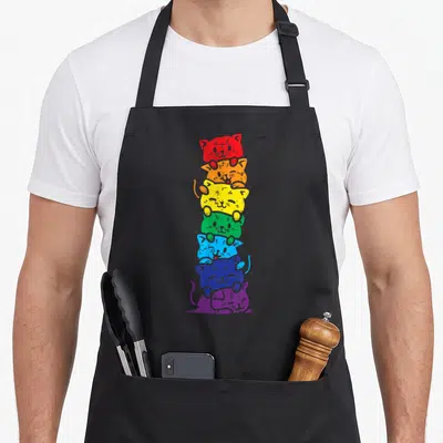 Cat rainbow apron - Seven cartoon cats stacked vertically in red orange yellow green blue and purple — shown on a black cotton canvas apron against a white background