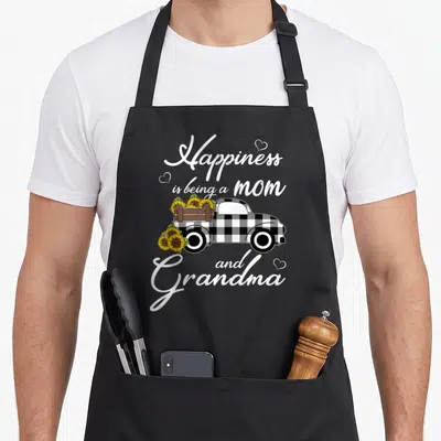 Happiness truck apron - Buffalo plaid truck carrying sunflowers with white script text and hearts — shown on a black cotton canvas apron against a white background