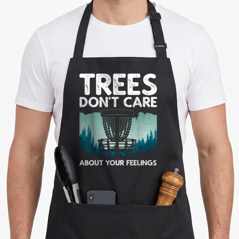 Disc player golf apron - Distressed disc golf basket with Trees Don't Care About Your Feelings text over forest — shown on a black cotton canvas apron against a white background