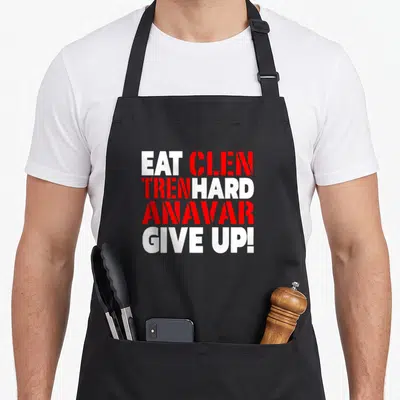 Eat hard quotes apron - Stacked white and red text reading EAT CLEN TREN HARD ANAVAR GIVE UP on black background — shown on a black cotton canvas apron against a white background
