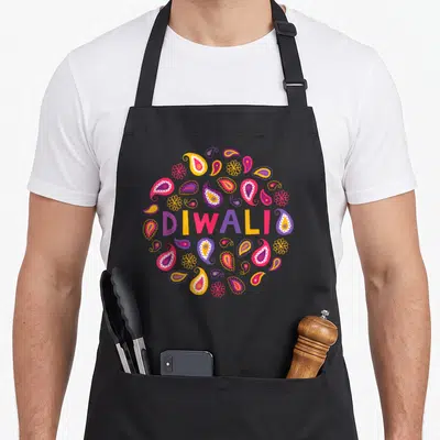 Funny diwali vibes apron - DIWALI text in pink yellow purple orange surrounded by circular paisley and flower patterns — shown on a black cotton canvas apron against a white background
