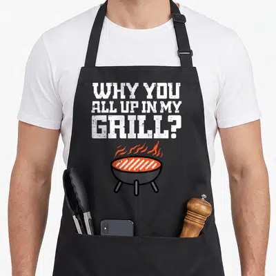 Funny barbecue apron - WHY YOU ALL UP IN MY GRILL text above cartoon charcoal grill with orange flames on black fabric — shown on a black cotton canvas apron against a white background