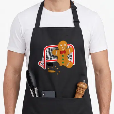 Hockey gingerbread apron - Cartoon gingerbread man goalie with red bow tie in front of hockey net and puck — shown on a black cotton canvas apron against a white background