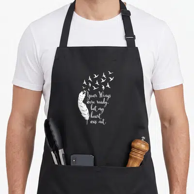 Wing memory apron - White feather and flock of birds with memorial quote in cursive script — shown on a black cotton canvas apron against a white background