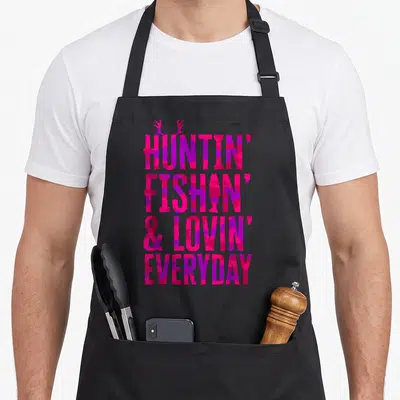 Fish camo apron - Pink purple camo text HUNTIN FISHIN LOVIN EVERYDAY with antlers and fish silhouette — shown on a black cotton canvas apron against a white background